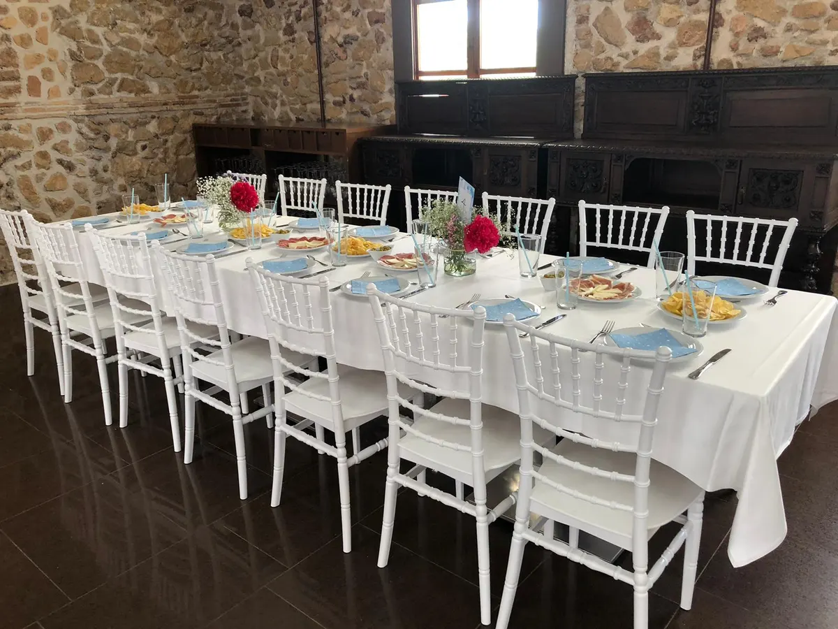 Mesa larga de celebración con sillas blancas, flores y aperitivos, ante un muro de piedra y aparadores tallados