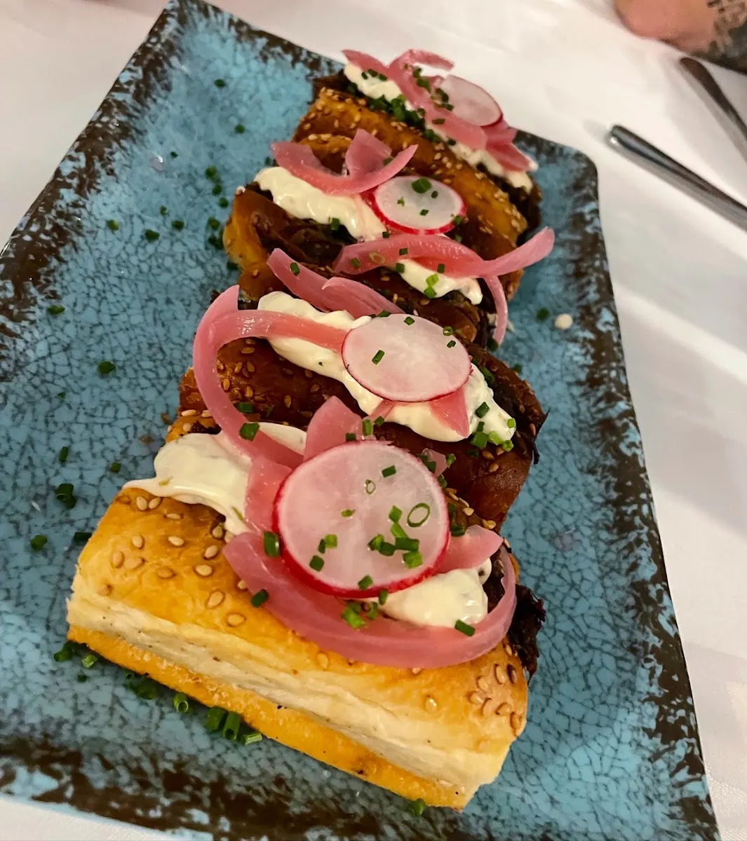 Pieza de masa dorada con sésamo, rellena y coronada con salsa blanca, rabanito, cebolla encurtida y cebollino, en fuente azul