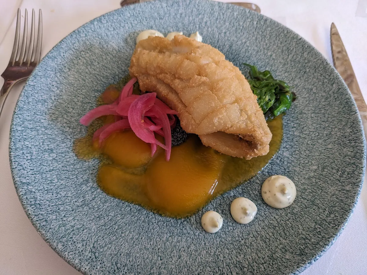 Pescado rebozado sobre crema anaranjada, con cebolla encurtida, alga y puntos de mayonesa, en plato azul moteado
