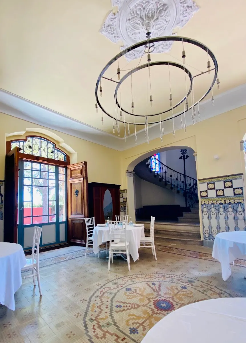 Salón de la villa con suelo de mosaico, zócalo de azulejo, lámpara de aros, puerta acristalada en arco y la escalera de forja al fondo